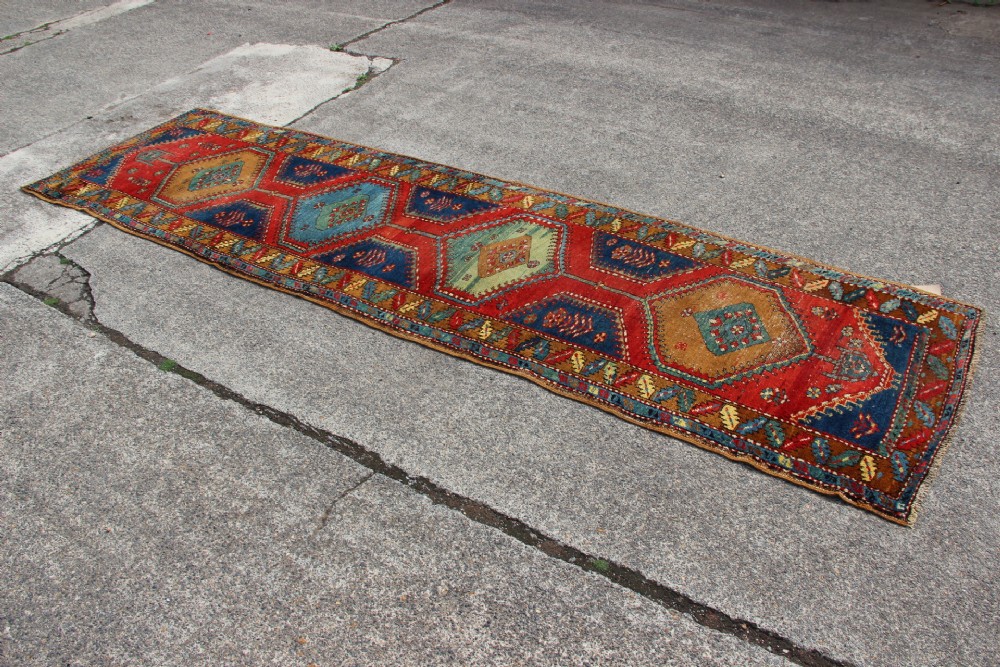An antique North West Persian long hallway runner rug with a red and blue field, featuring geometric diamond medallions and a stylized leaf border. View IV