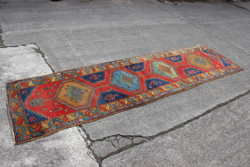 An antique North West Persian long hallway runner rug with a red and blue field, featuring geometric diamond medallions and a stylized leaf border. View V