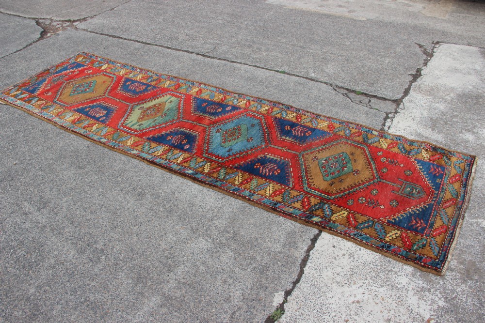 An antique North West Persian long hallway runner rug with a red and blue field, featuring geometric diamond medallions and a stylized leaf border. View VI