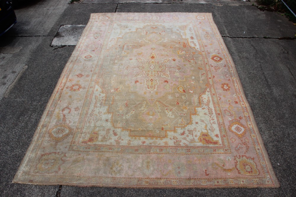 A large hand-knotted wool Oushak carpet displaying a central star medallion, angular floral spandrels, and a pastel palette of pale pink, cream, and beige.