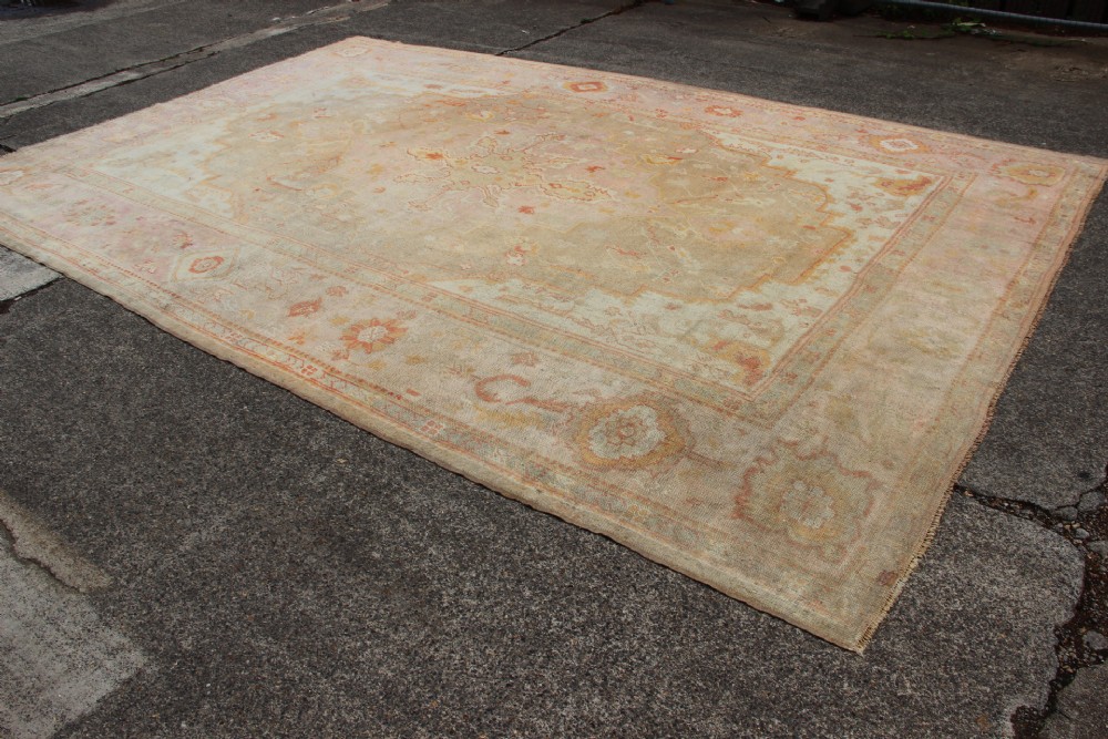 A large hand-knotted wool Oushak carpet displaying a central star medallion, angular floral spandrels, and a pastel palette of pale pink, cream, and beige. View XII