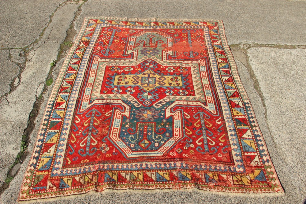 A hand-knotted Caucasian rug featuring a central red and blue shield medallion framed by geometric borders on a red field.