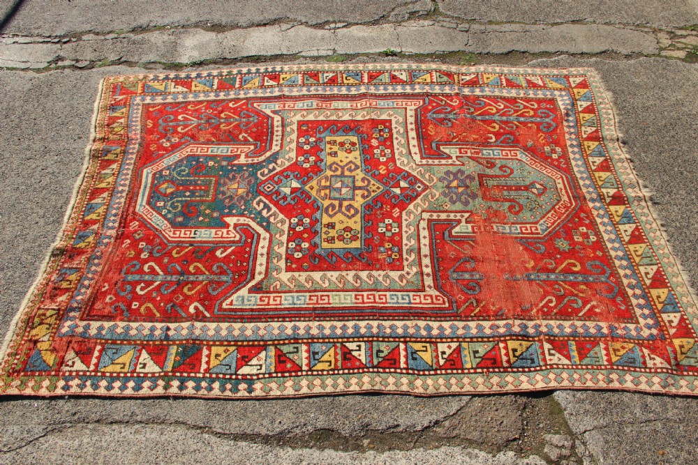 A hand-knotted Caucasian rug featuring a central red and blue shield medallion framed by geometric borders on a red field. View III