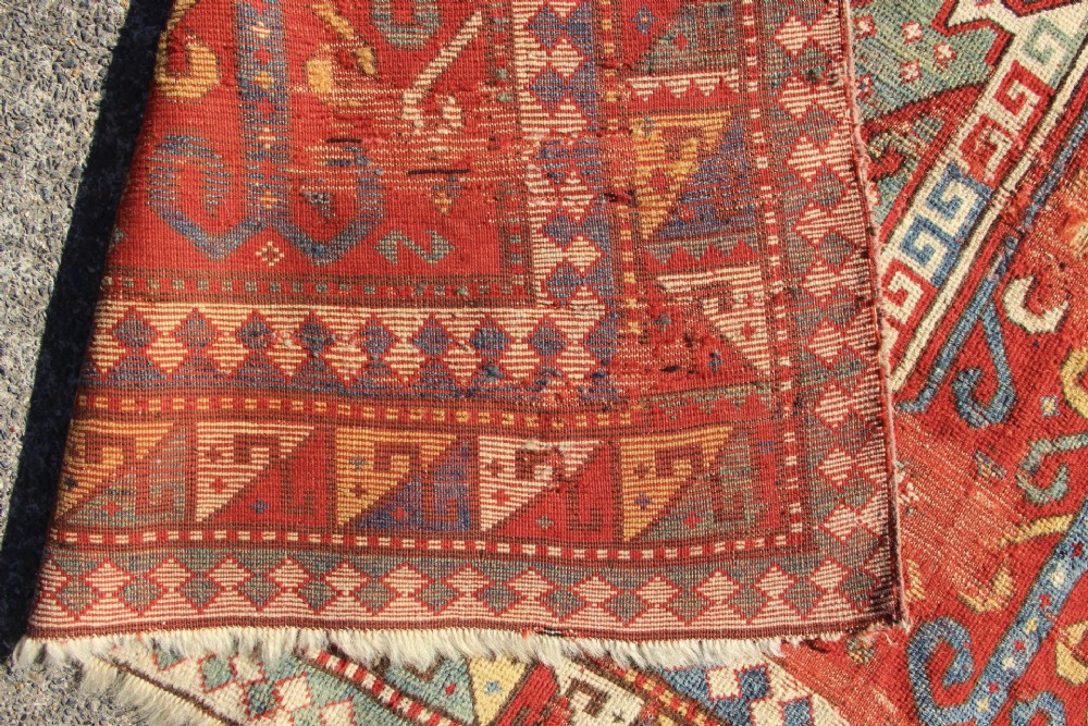 A hand-knotted Caucasian rug featuring a central red and blue shield medallion framed by geometric borders on a red field. View IV
