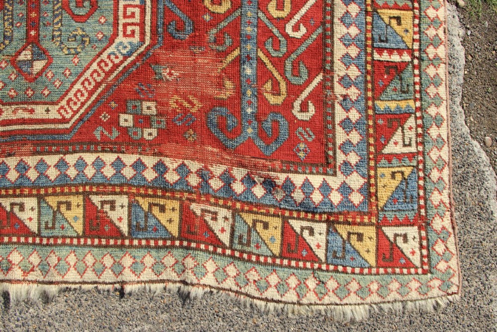 A hand-knotted Caucasian rug featuring a central red and blue shield medallion framed by geometric borders on a red field. View V