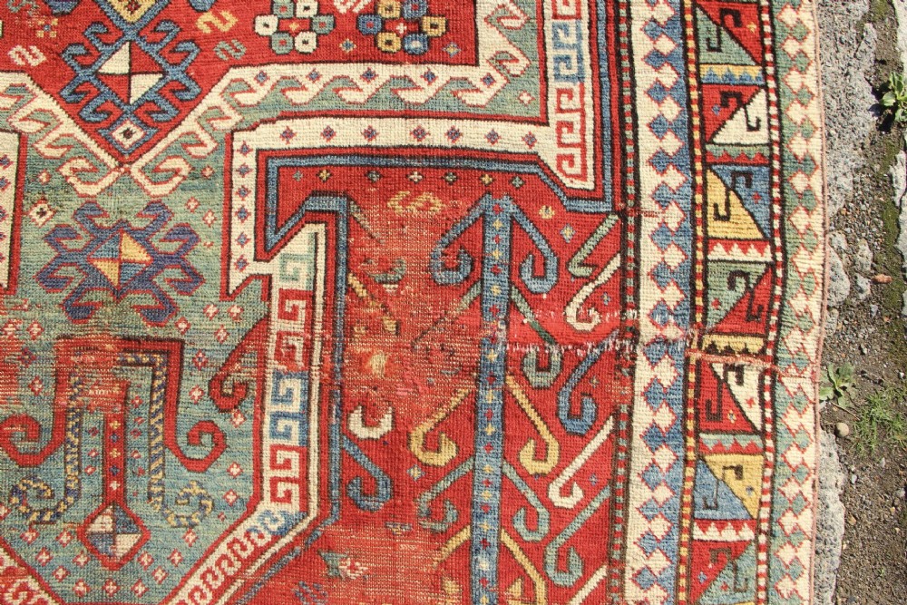 A hand-knotted Caucasian rug featuring a central red and blue shield medallion framed by geometric borders on a red field. View VI