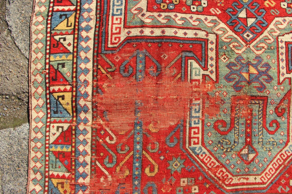 A hand-knotted Caucasian rug featuring a central red and blue shield medallion framed by geometric borders on a red field. View VII