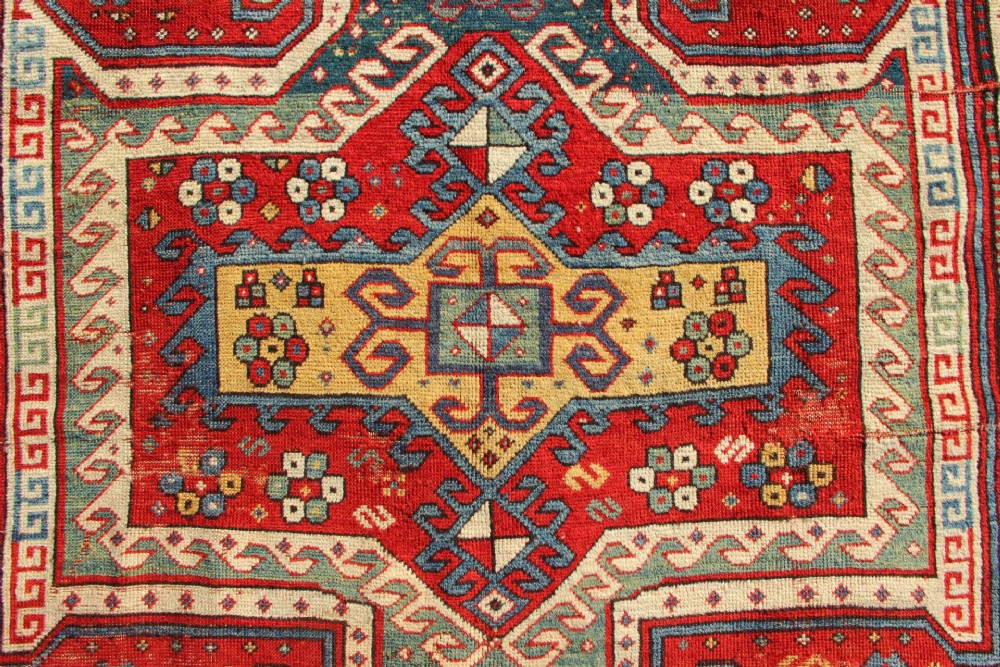 A hand-knotted Caucasian rug featuring a central red and blue shield medallion framed by geometric borders on a red field. View VIII