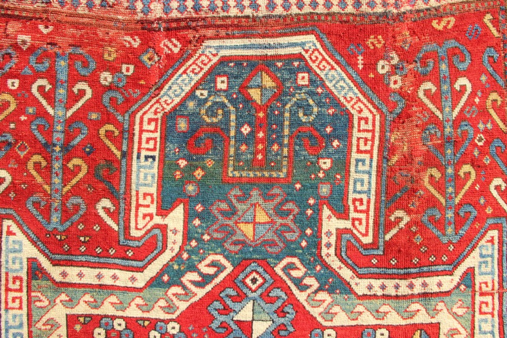 A hand-knotted Caucasian rug featuring a central red and blue shield medallion framed by geometric borders on a red field. View IX
