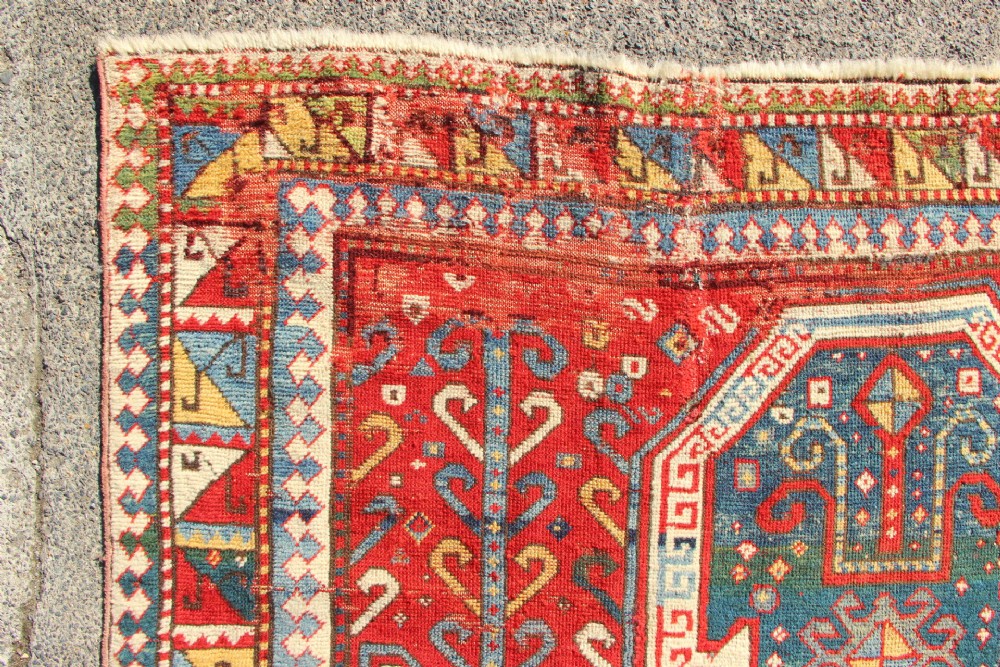 A hand-knotted Caucasian rug featuring a central red and blue shield medallion framed by geometric borders on a red field. View X