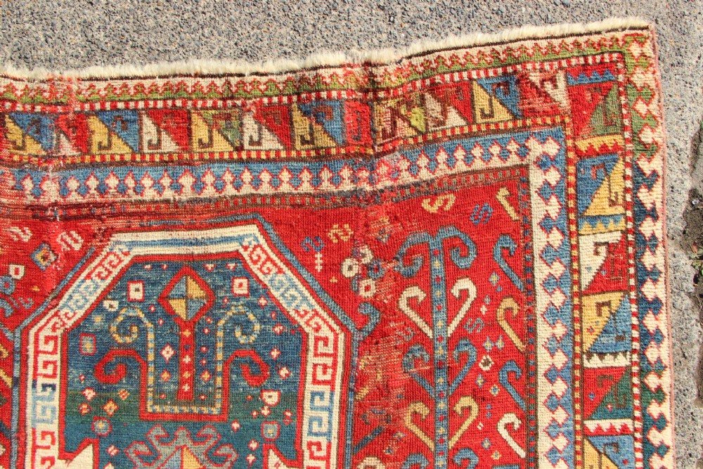 A hand-knotted Caucasian rug featuring a central red and blue shield medallion framed by geometric borders on a red field. View XI