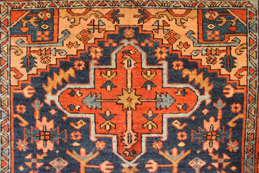 An antique hand-knotted Heriz wool rug featuring a geometric central medallion surrounded by intricate tribal patterns in terracotta red, navy blue, and cream tones. View IV