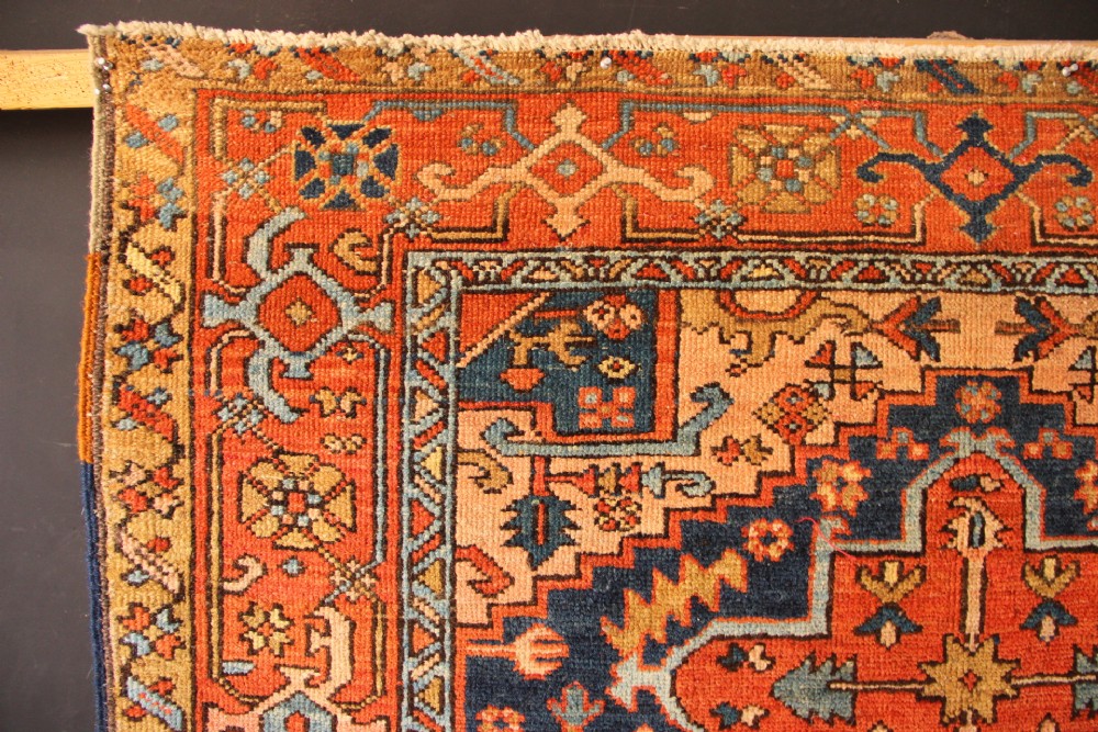 An antique hand-knotted Heriz wool rug featuring a geometric central medallion surrounded by intricate tribal patterns in terracotta red, navy blue, and cream tones. View V