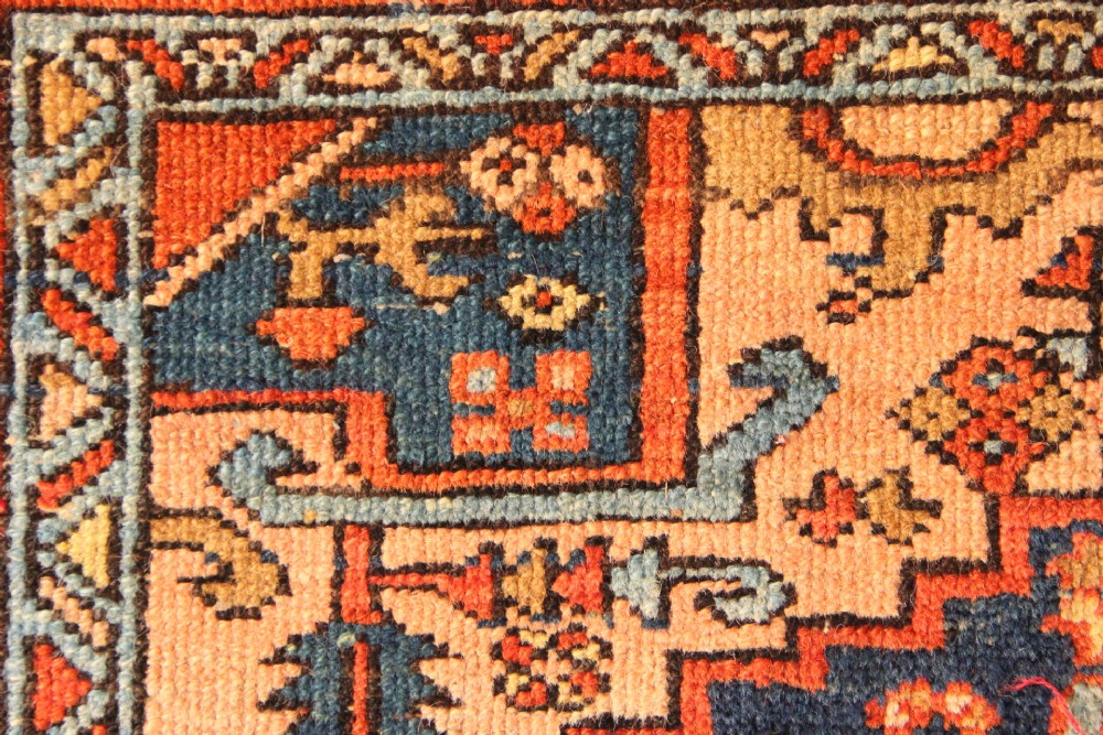 An antique hand-knotted Heriz wool rug featuring a geometric central medallion surrounded by intricate tribal patterns in terracotta red, navy blue, and cream tones. View VI