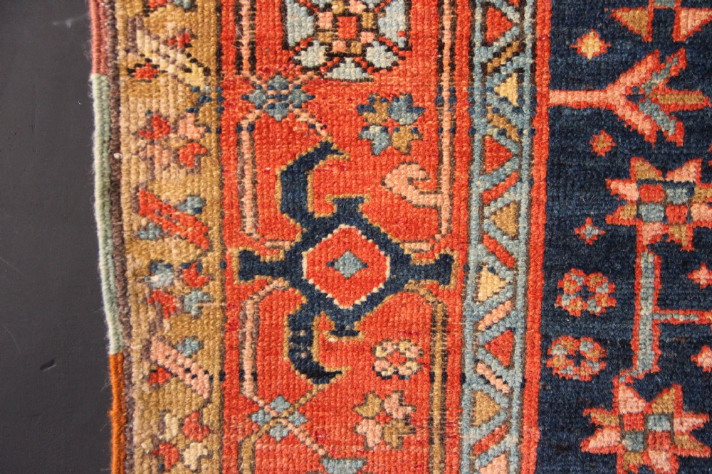 An antique hand-knotted Heriz wool rug featuring a geometric central medallion surrounded by intricate tribal patterns in terracotta red, navy blue, and cream tones. View VII