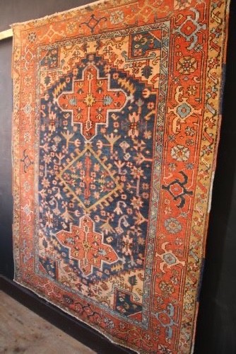 An antique hand-knotted Heriz wool rug featuring a geometric central medallion surrounded by intricate tribal patterns in terracotta red, navy blue, and cream tones. View X