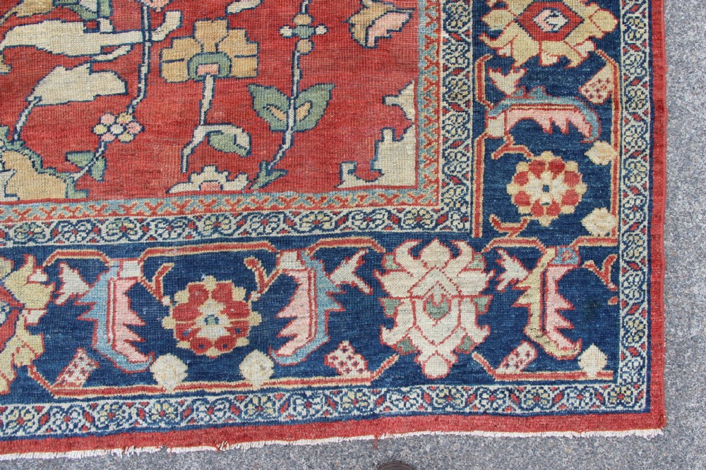 A large hand-knotted wool carpet featuring a terracotta central field with scrolling beige floral motifs framed by a wide navy blue patterned border. View II