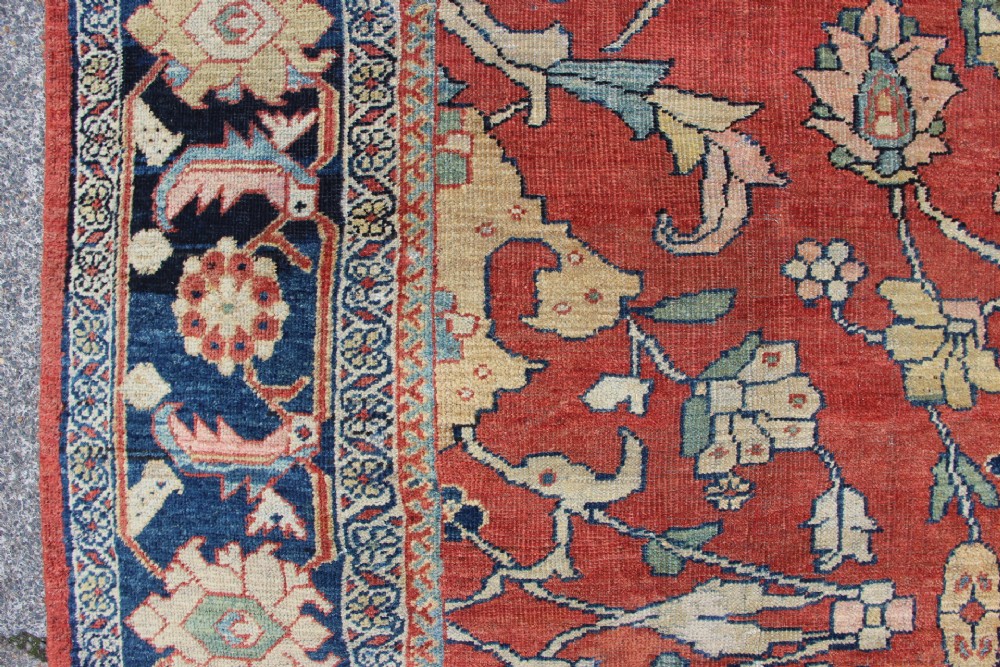A large hand-knotted wool carpet featuring a terracotta central field with scrolling beige floral motifs framed by a wide navy blue patterned border. View IV