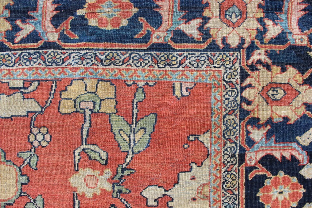 A large hand-knotted wool carpet featuring a terracotta central field with scrolling beige floral motifs framed by a wide navy blue patterned border. View VII