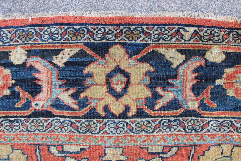 A large hand-knotted wool carpet featuring a terracotta central field with scrolling beige floral motifs framed by a wide navy blue patterned border. View VIII