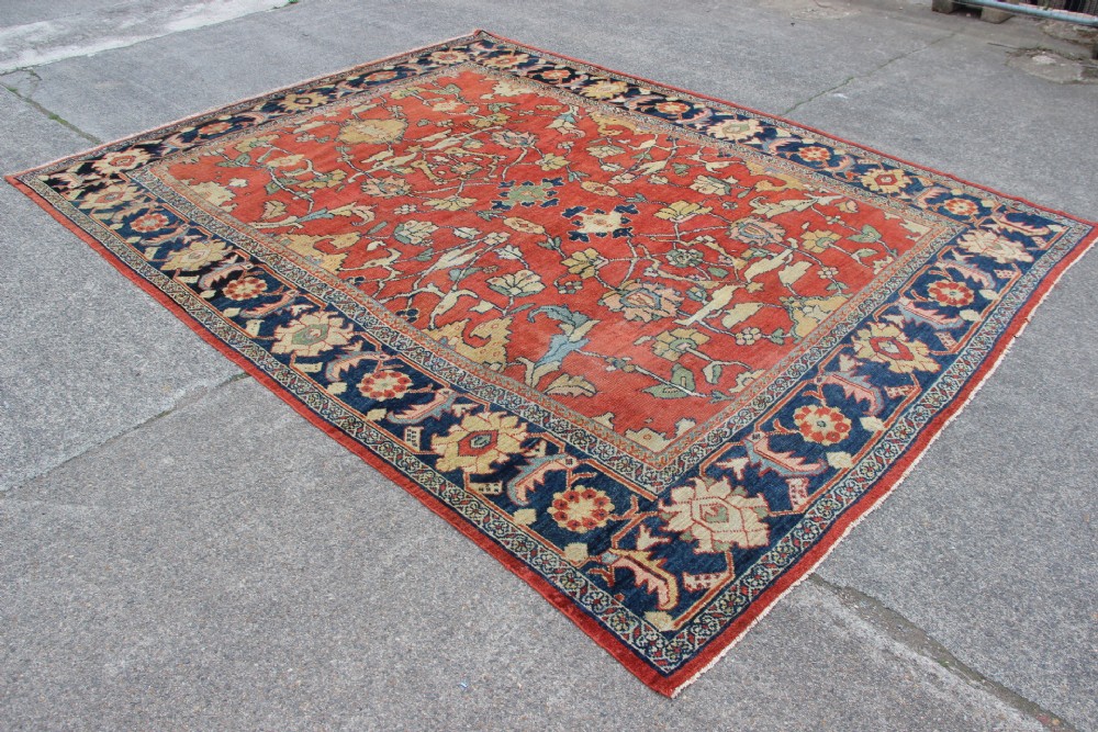 A large hand-knotted wool carpet featuring a terracotta central field with scrolling beige floral motifs framed by a wide navy blue patterned border. View IX