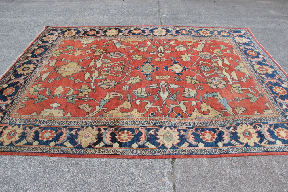 A large hand-knotted wool carpet featuring a terracotta central field with scrolling beige floral motifs framed by a wide navy blue patterned border. View X