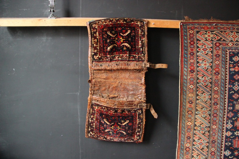 An antique miniature Khamseh tribal saddlebag featuring two hand-woven wool panels with deep red and navy geometric bird motifs joined by a leather centre section with straps.