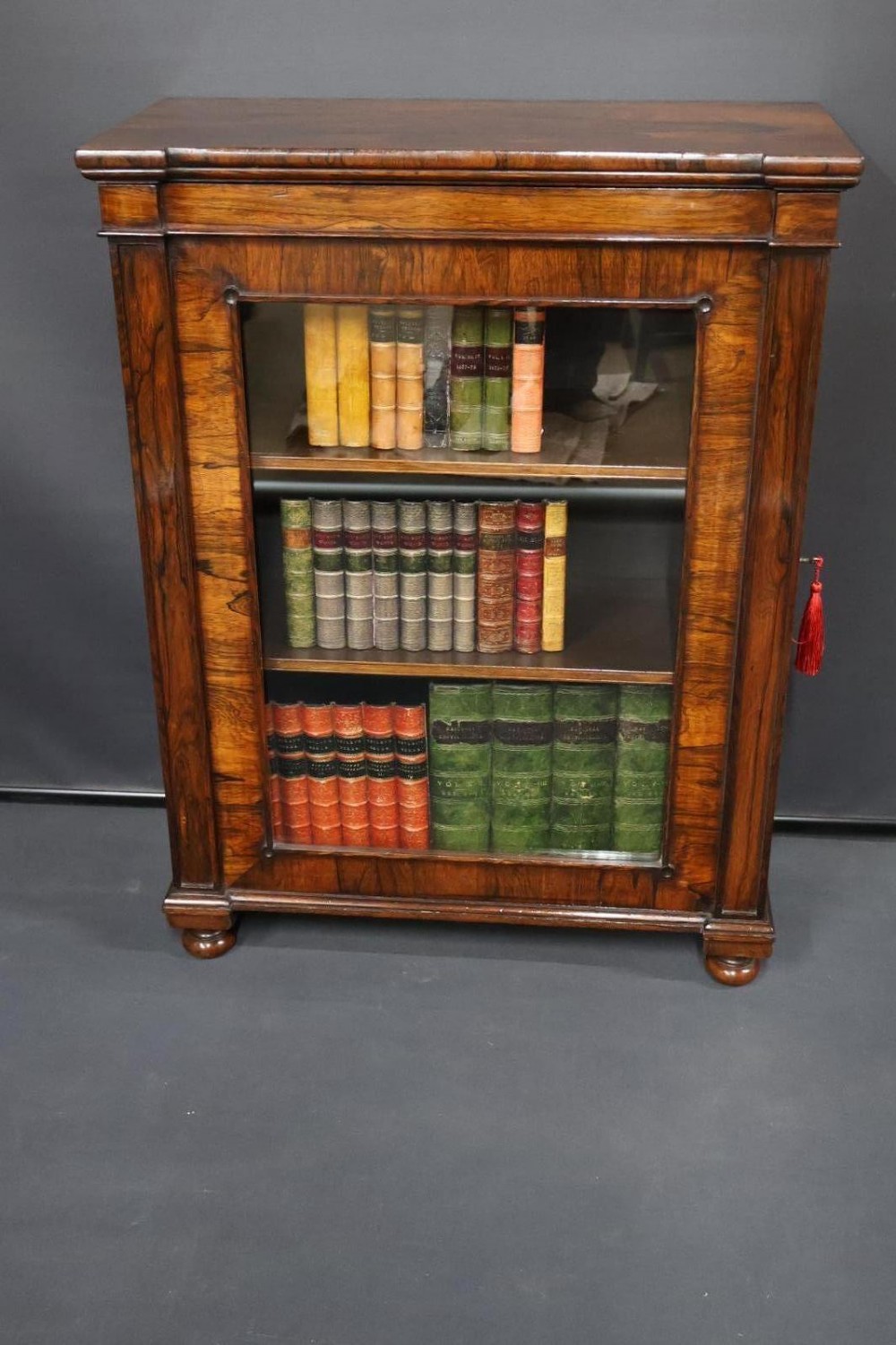 A Regency dark rosewood pier cabinet featuring a single glazed door revealing internal shelves, set on turned bun feet.
