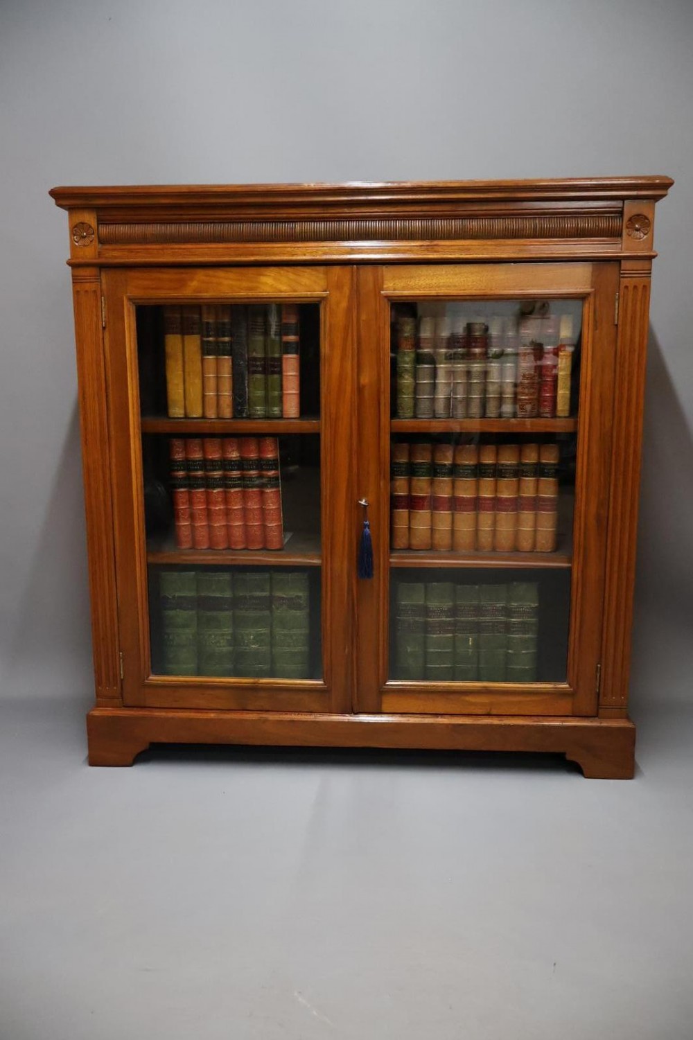 An Edwardian low walnut bookcase featuring two glass-fronted doors, reeded frieze detailing, fluted pilasters, and shelves filled with leather-bound books.