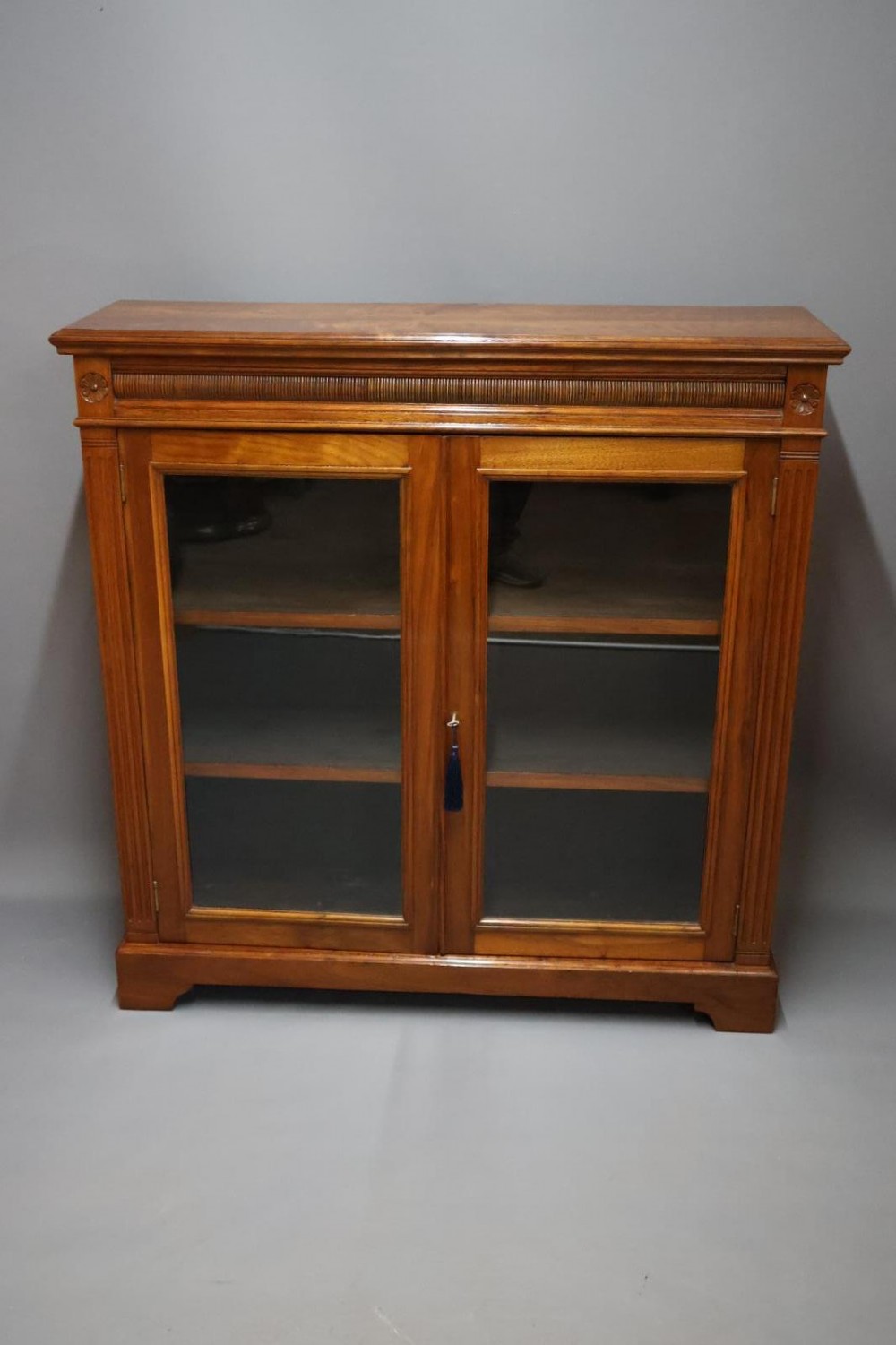 An Edwardian low walnut bookcase featuring two glass-fronted doors, reeded frieze detailing, fluted pilasters, and shelves filled with leather-bound books. View II