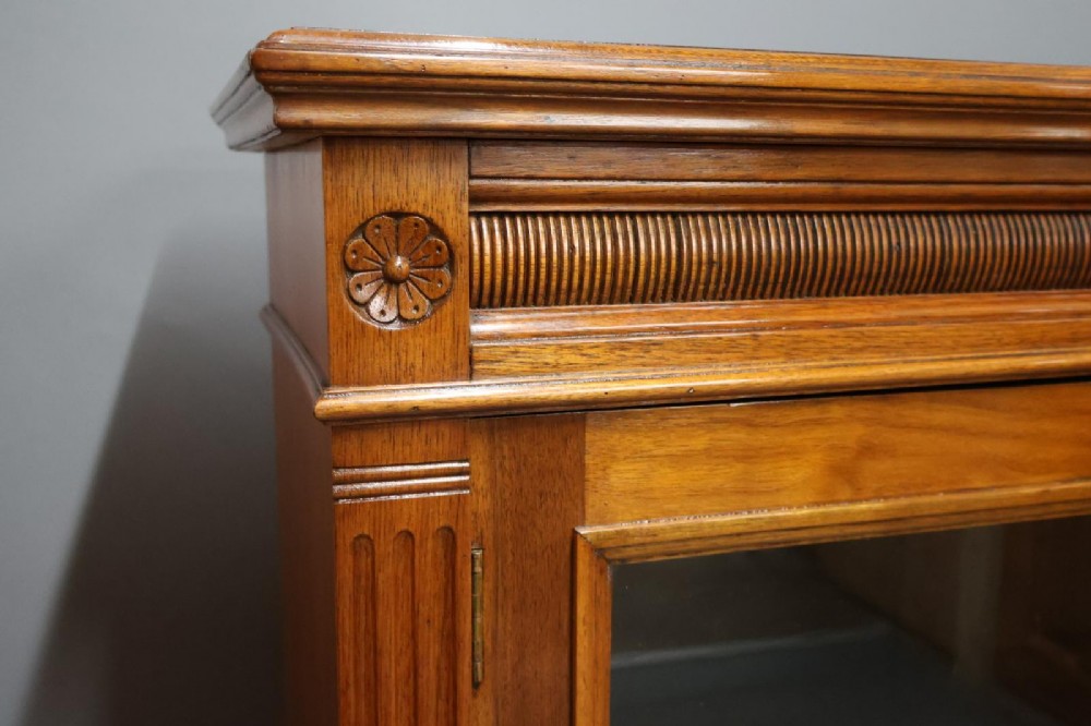 An Edwardian low walnut bookcase featuring two glass-fronted doors, reeded frieze detailing, fluted pilasters, and shelves filled with leather-bound books. View III
