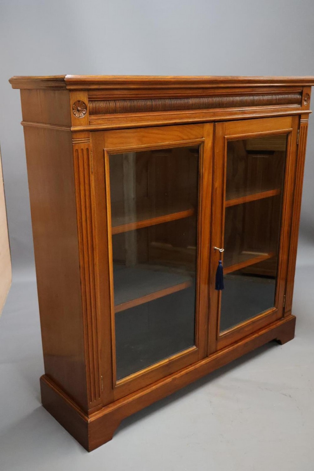 An Edwardian low walnut bookcase featuring two glass-fronted doors, reeded frieze detailing, fluted pilasters, and shelves filled with leather-bound books. View IV