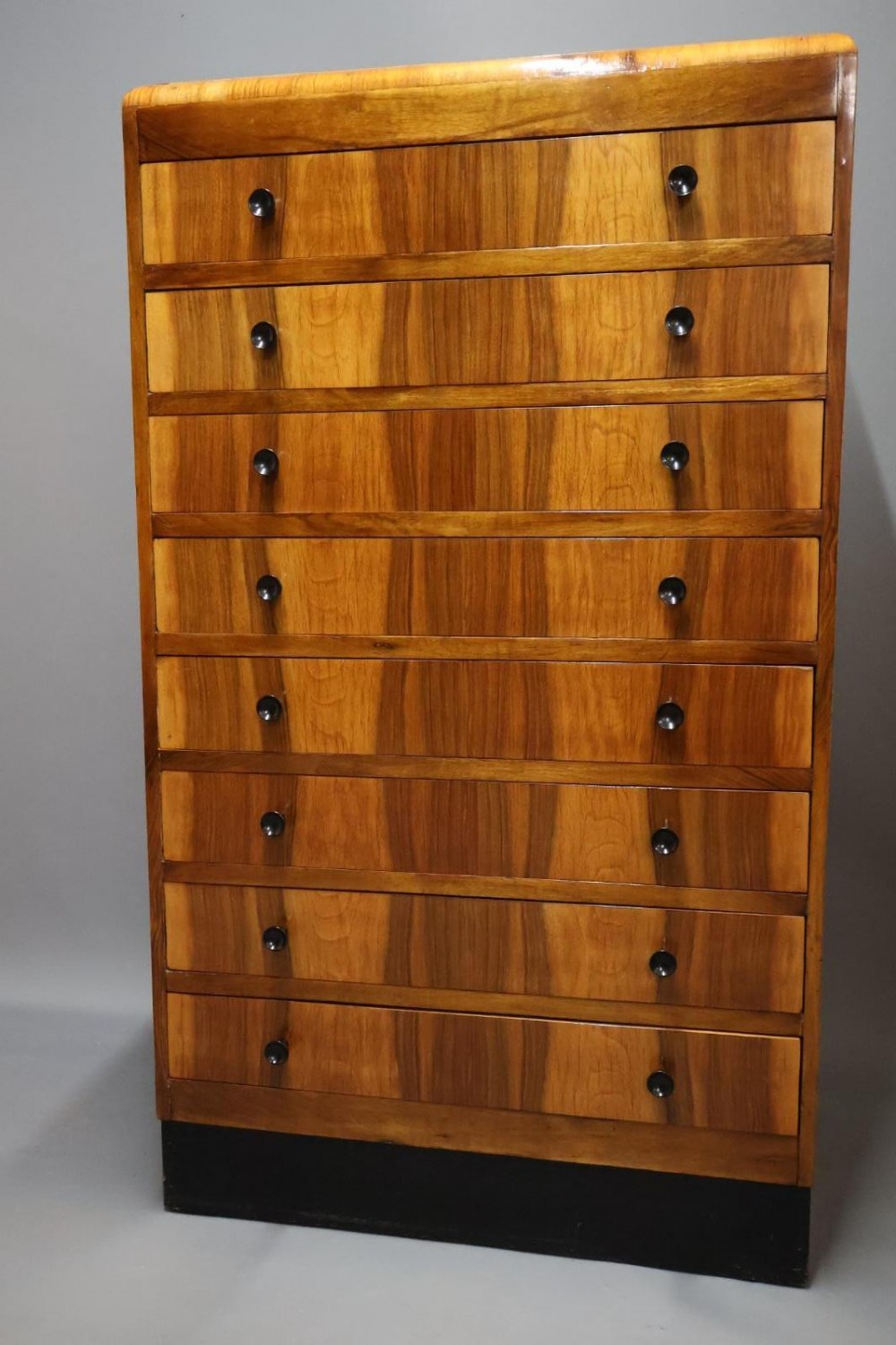 An Art Deco walnut tallboy chest of drawers featuring eight vertically stacked drawers with figured wood grain, round black handles, and an ebonised base.