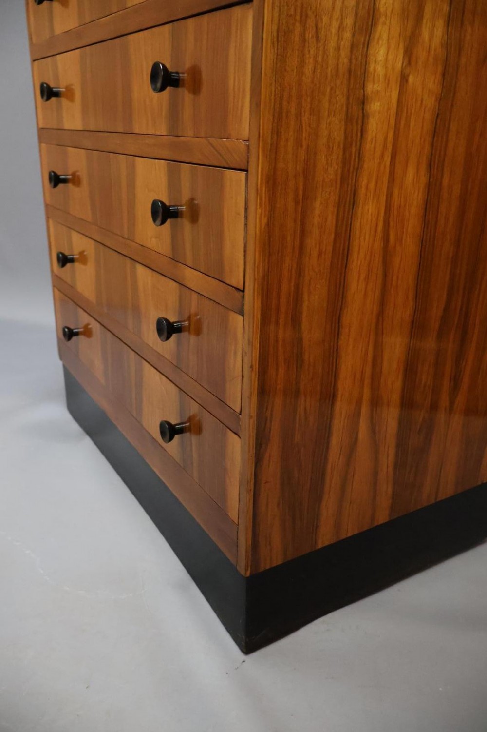An Art Deco walnut tallboy chest of drawers featuring eight vertically stacked drawers with figured wood grain, round black handles, and an ebonised base. View III