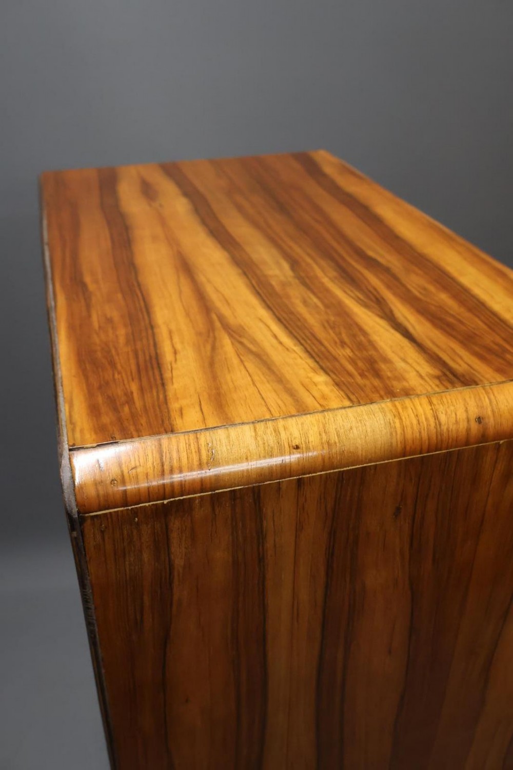 An Art Deco walnut tallboy chest of drawers featuring eight vertically stacked drawers with figured wood grain, round black handles, and an ebonised base. View IV