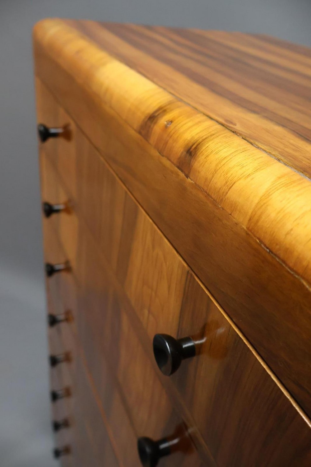 An Art Deco walnut tallboy chest of drawers featuring eight vertically stacked drawers with figured wood grain, round black handles, and an ebonised base. View V