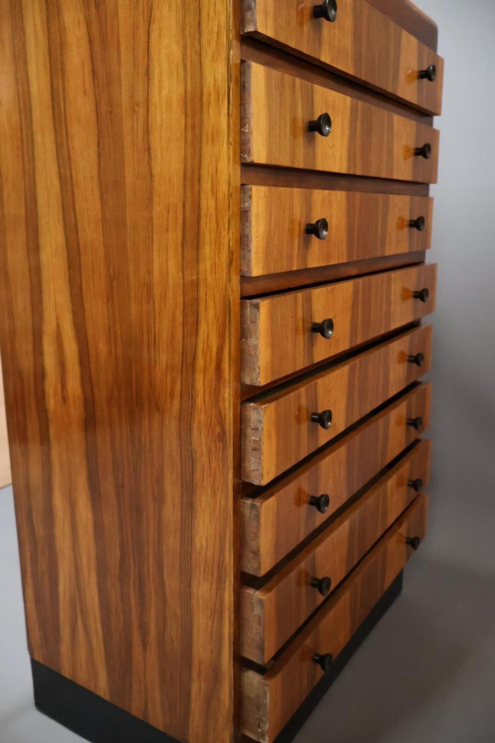 An Art Deco walnut tallboy chest of drawers featuring eight vertically stacked drawers with figured wood grain, round black handles, and an ebonised base. View VI