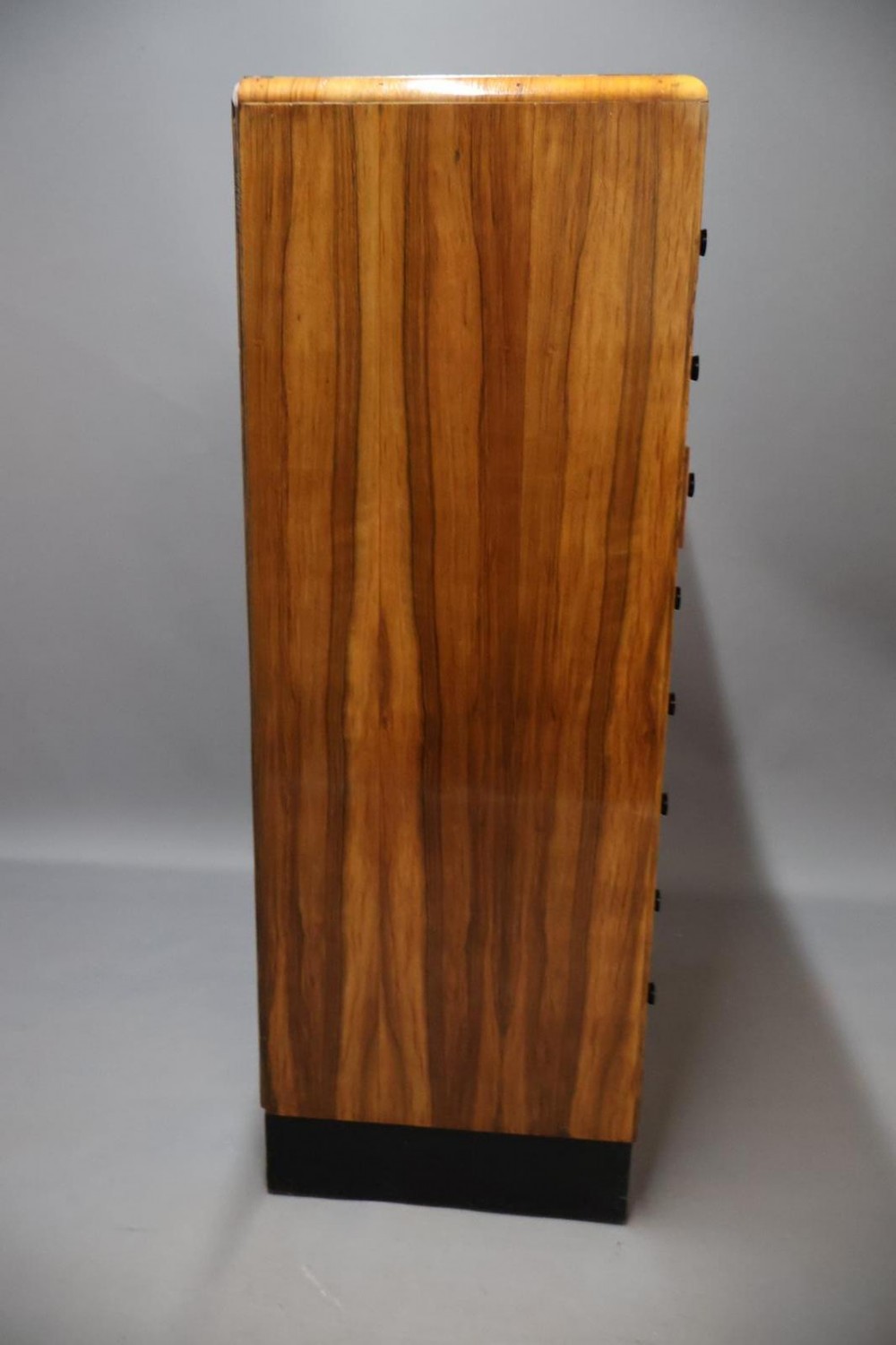 An Art Deco walnut tallboy chest of drawers featuring eight vertically stacked drawers with figured wood grain, round black handles, and an ebonised base. View VII