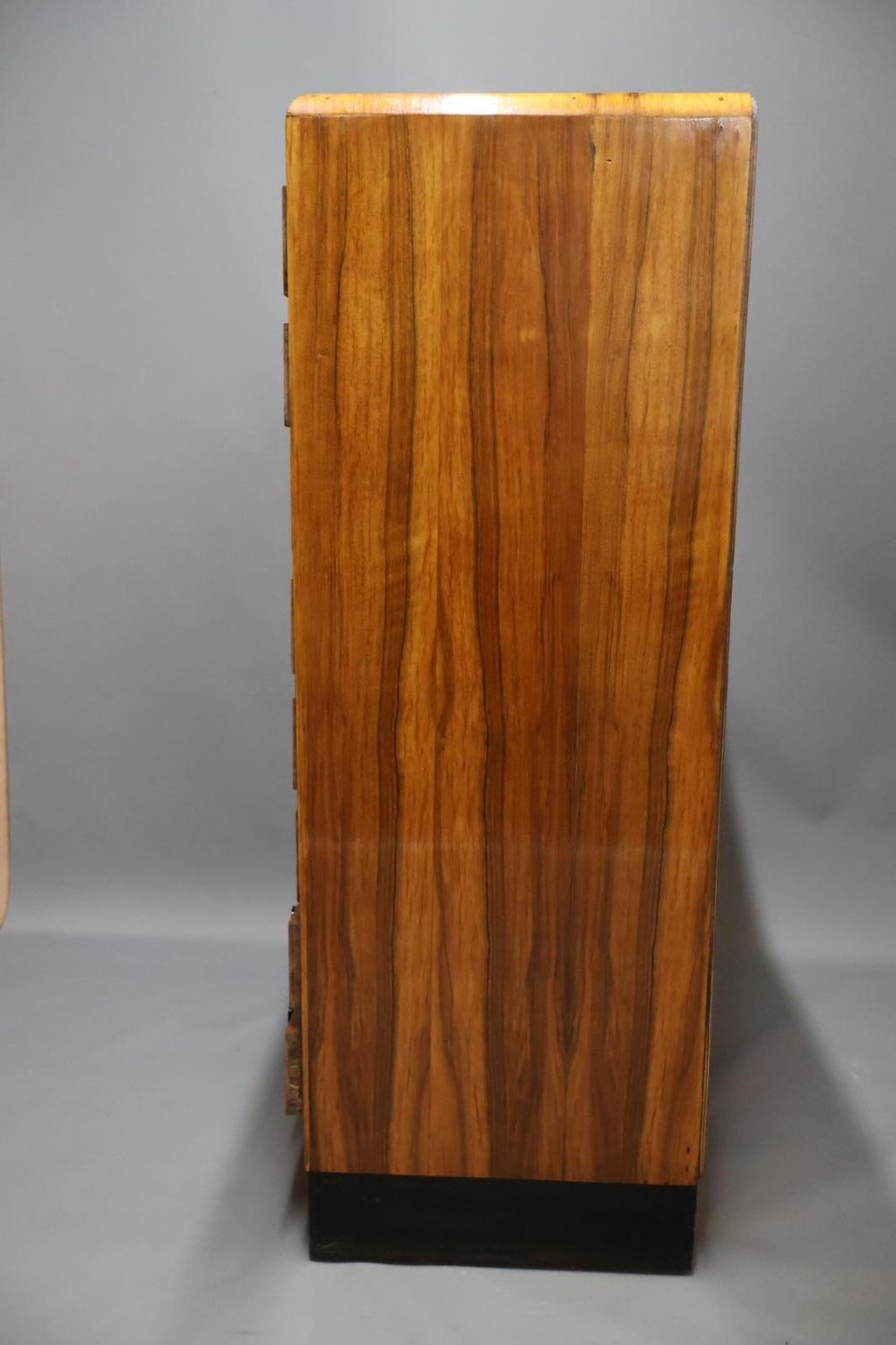 An Art Deco walnut tallboy chest of drawers featuring eight vertically stacked drawers with figured wood grain, round black handles, and an ebonised base. View IX