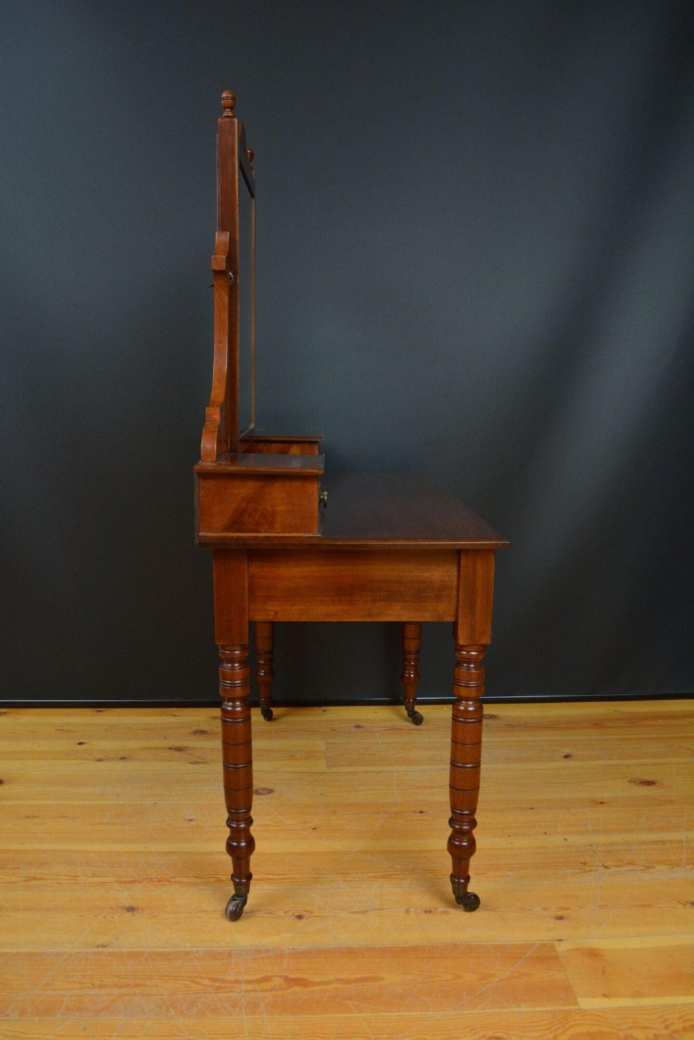 An Edwardian mahogany dressing table featuring a central rectangular mirror flanked by small jewellery drawers, resting on turned legs with brass casters. View VII