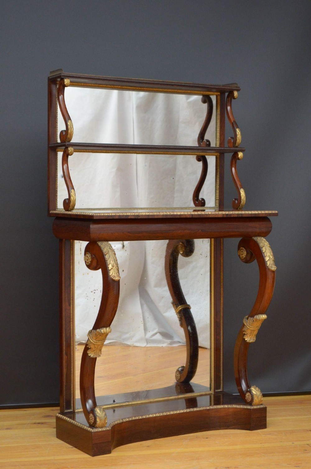 A Regency rosewood console table featuring an upper shelf tier, mirror-backed panels, and gilt carved scroll legs. View II