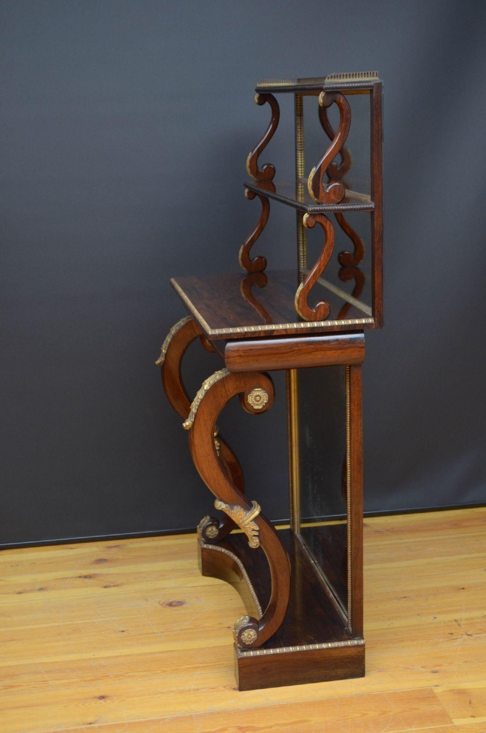 A Regency rosewood console table featuring an upper shelf tier, mirror-backed panels, and gilt carved scroll legs. View X