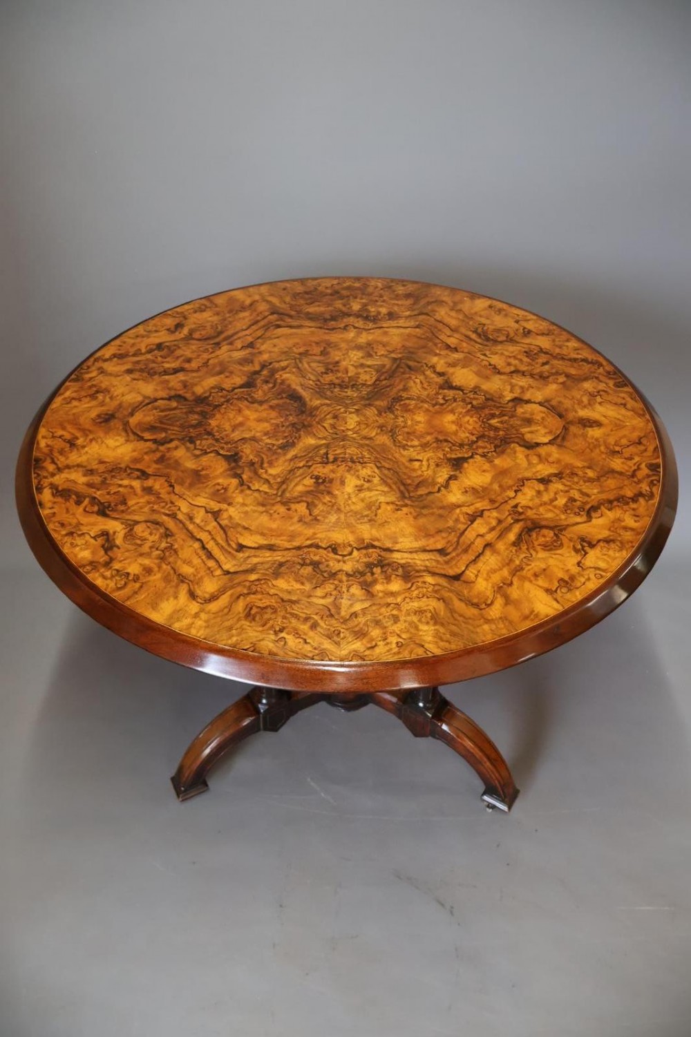 A circular burr walnut centre table featuring a figured top, turned central pedestal, four outer colonnettes, and splayed legs on castors. View II