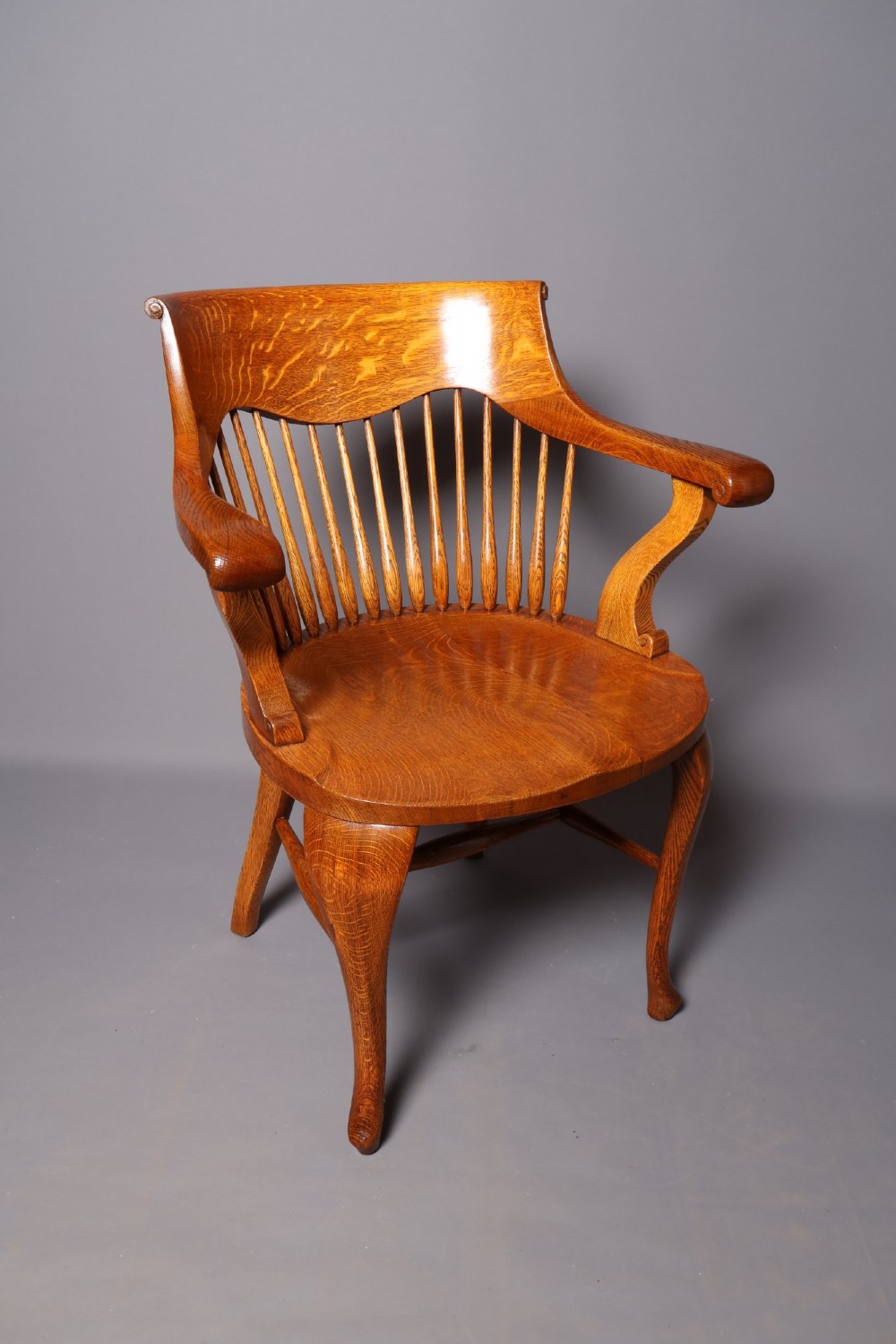An antique Victorian golden oak desk chair featuring a curved spindle back, curved armrests, a contoured seat, and cabriole front legs.