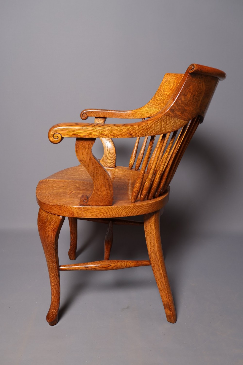 An antique Victorian golden oak desk chair featuring a curved spindle back, curved armrests, a contoured seat, and cabriole front legs. View III