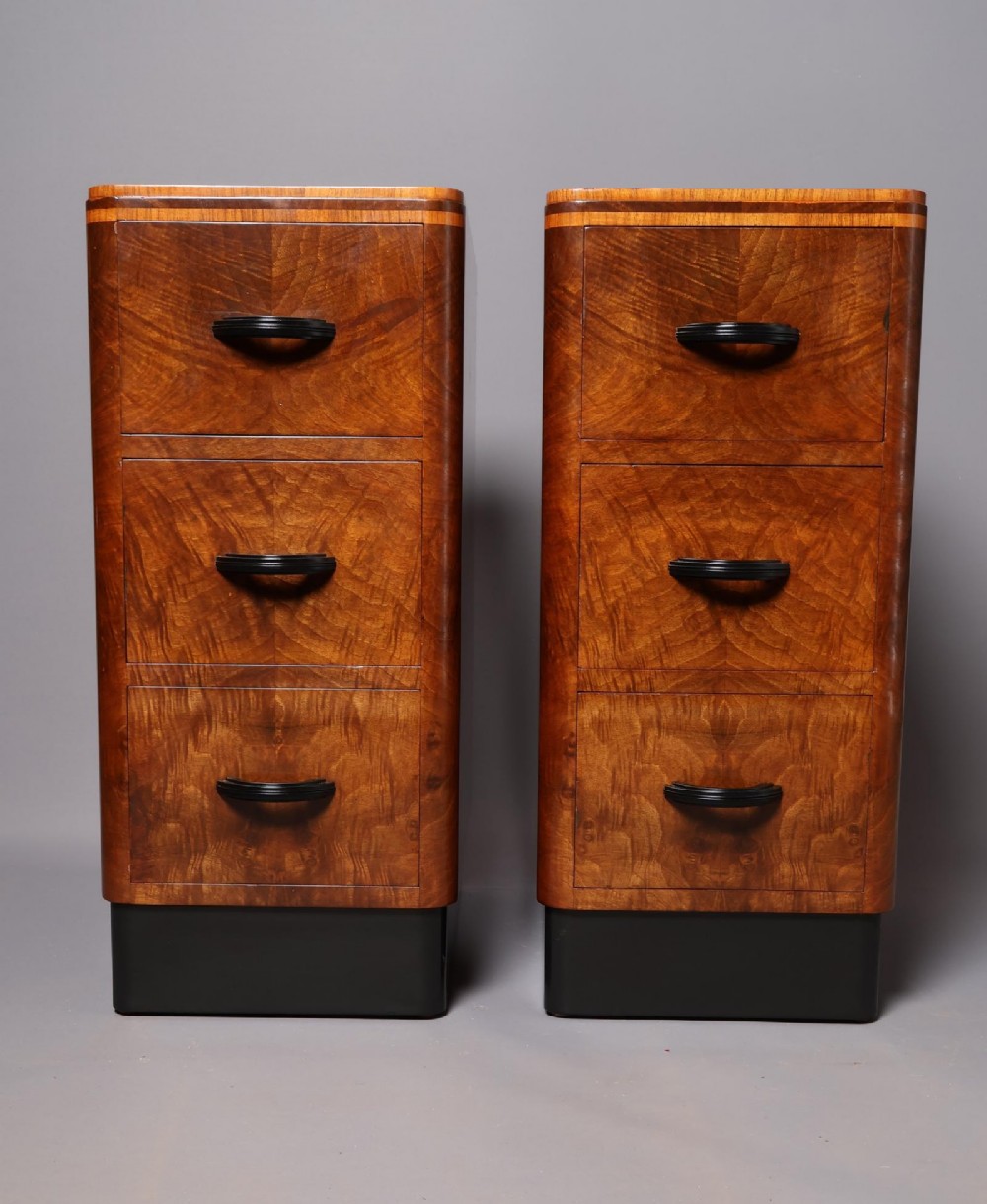 A matching pair of tall Art Deco burr walnut bedside cabinets with three drawers, dark curved handles, and ebonised plinth bases.