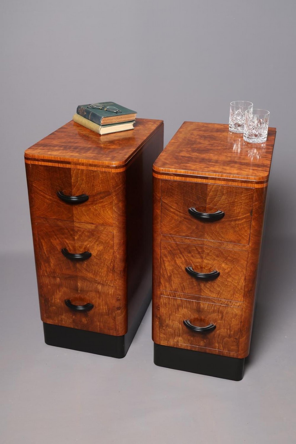 A matching pair of tall Art Deco burr walnut bedside cabinets with three drawers, dark curved handles, and ebonised plinth bases. View II