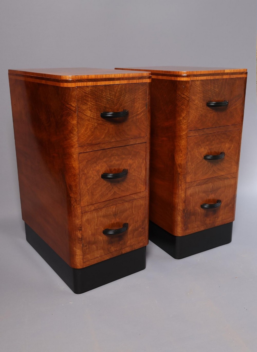 A matching pair of tall Art Deco burr walnut bedside cabinets with three drawers, dark curved handles, and ebonised plinth bases. View III