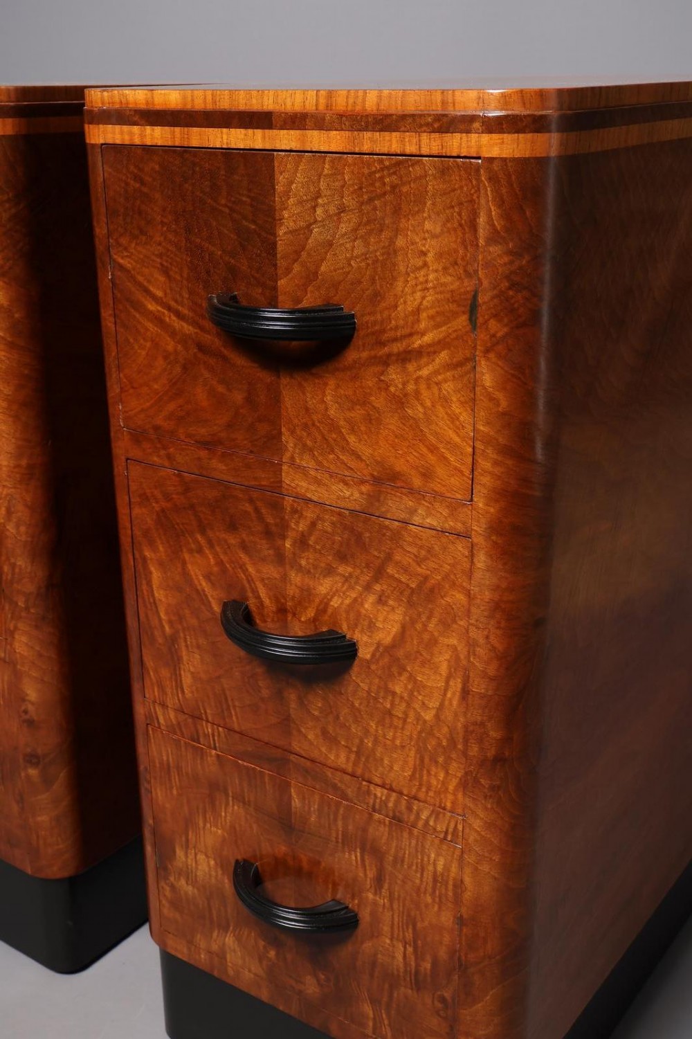 A matching pair of tall Art Deco burr walnut bedside cabinets with three drawers, dark curved handles, and ebonised plinth bases. View VI