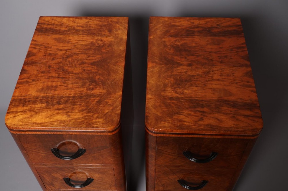 A matching pair of tall Art Deco burr walnut bedside cabinets with three drawers, dark curved handles, and ebonised plinth bases. View VII